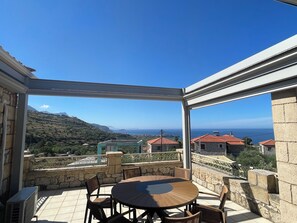 A covered outdoor patio with a round table and chairs, stone walls, and a view of a coastal landscape.