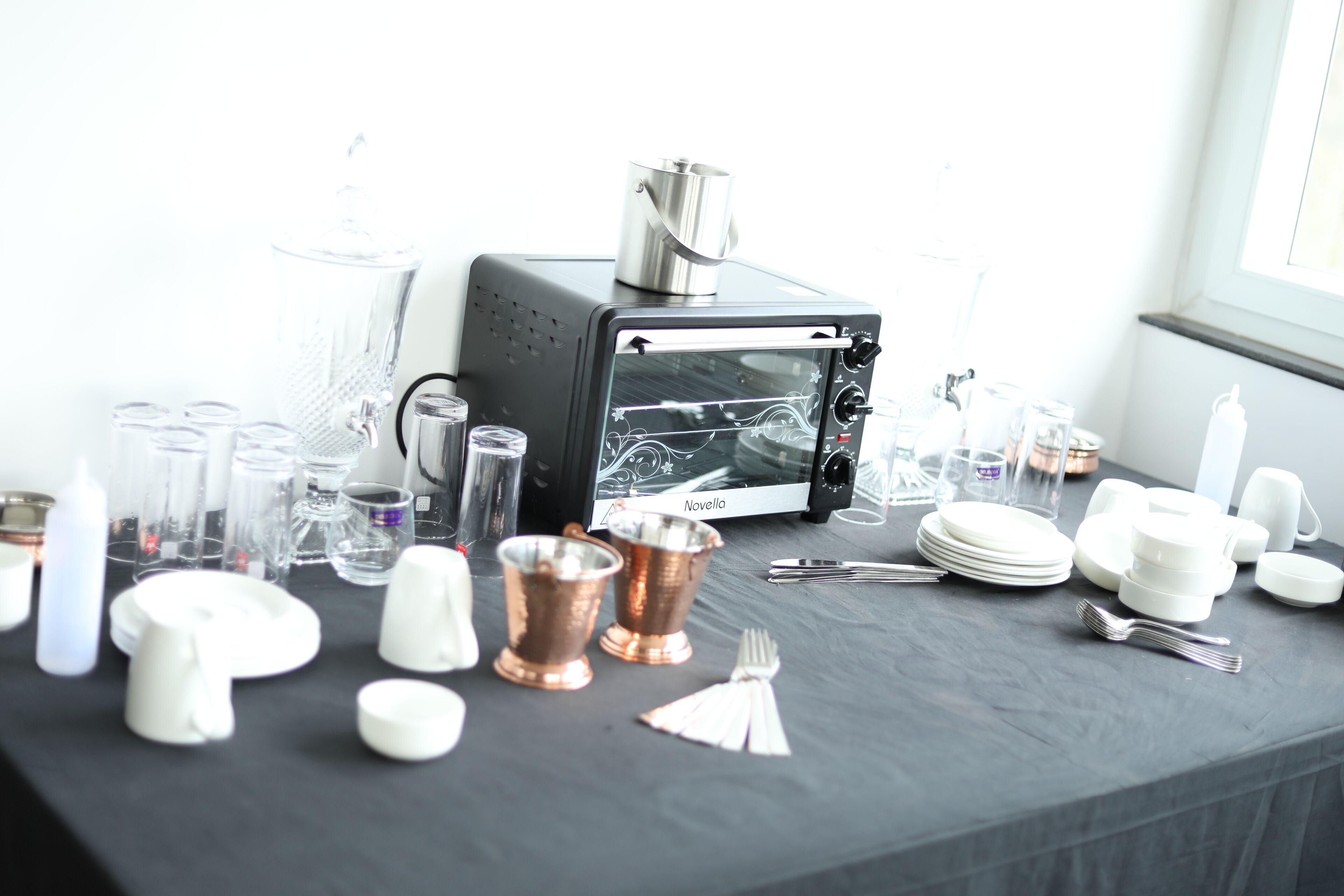 A table with a black tablecloth, a Novella toaster oven, and various glassware and dishes.