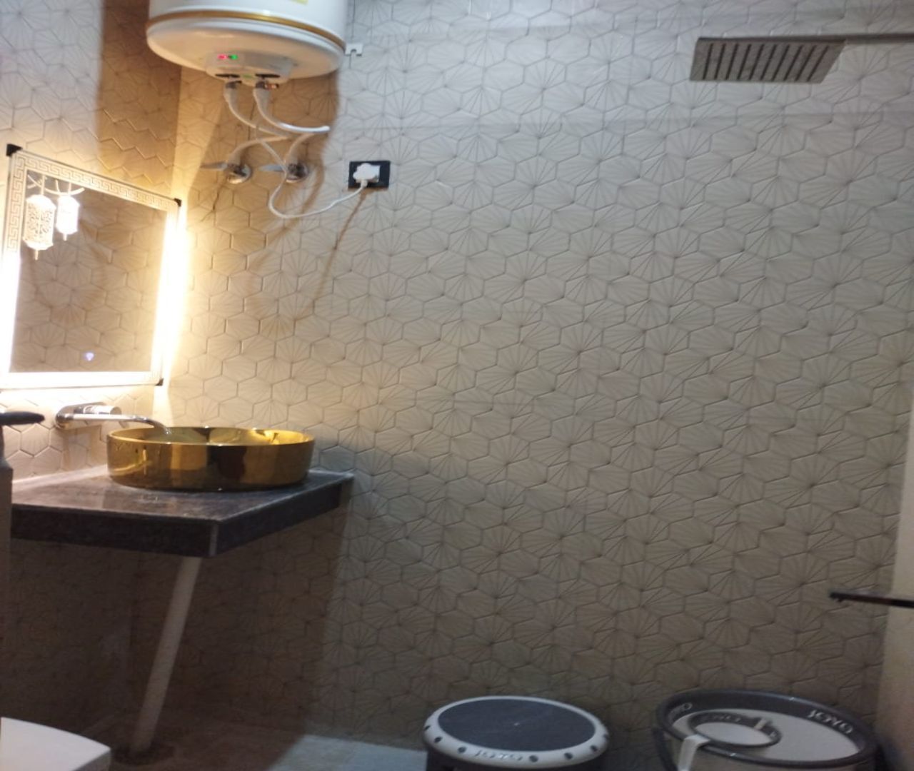 Une salle de bain avec un mur orné d’un motif hexagonal, un lavabo avec un receveur doré, des toilettes avec un abattant blanc et une petite poubelle.