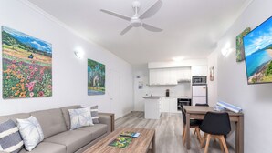 Ein modernes Wohnzimmer mit einer Couch, einem Couchtisch und einem Essbereich. Im Hintergrund ist eine Küche mit weißen Schränken und einer Mikrowelle zu sehen.