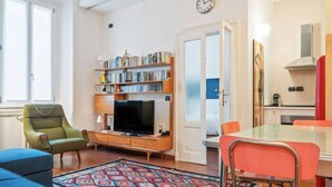 Una sala de estar con televisión, estantería, mesa de comedor, sillas y una alfombra estampada.