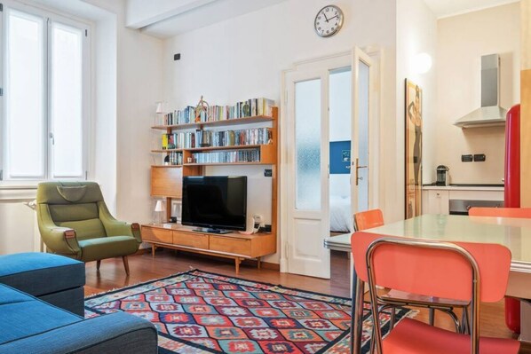 A living room with a TV, bookshelf, dining table, chairs, and a patterned rug.
