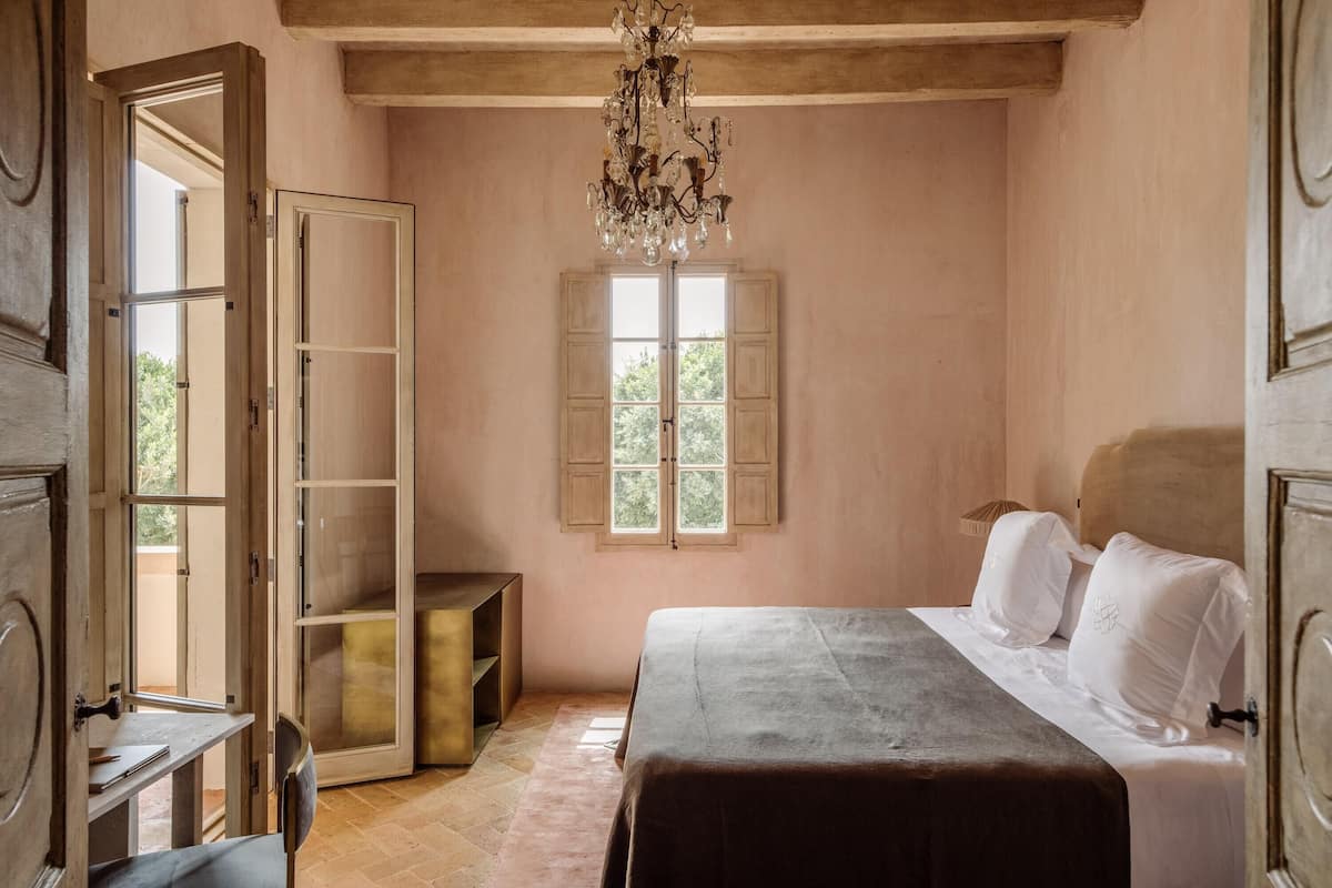 Un dormitorio con cama, candelabro, ventana con contraventanas y puerta de madera.