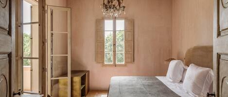 Un dormitorio con cama, candelabro, ventana con contraventanas y puerta de madera.