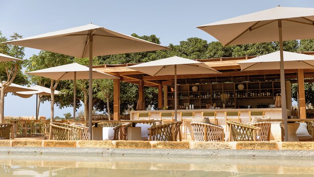 A beachside bar with outdoor seating under umbrellas, wooden chairs, and a bar counter with bottles.