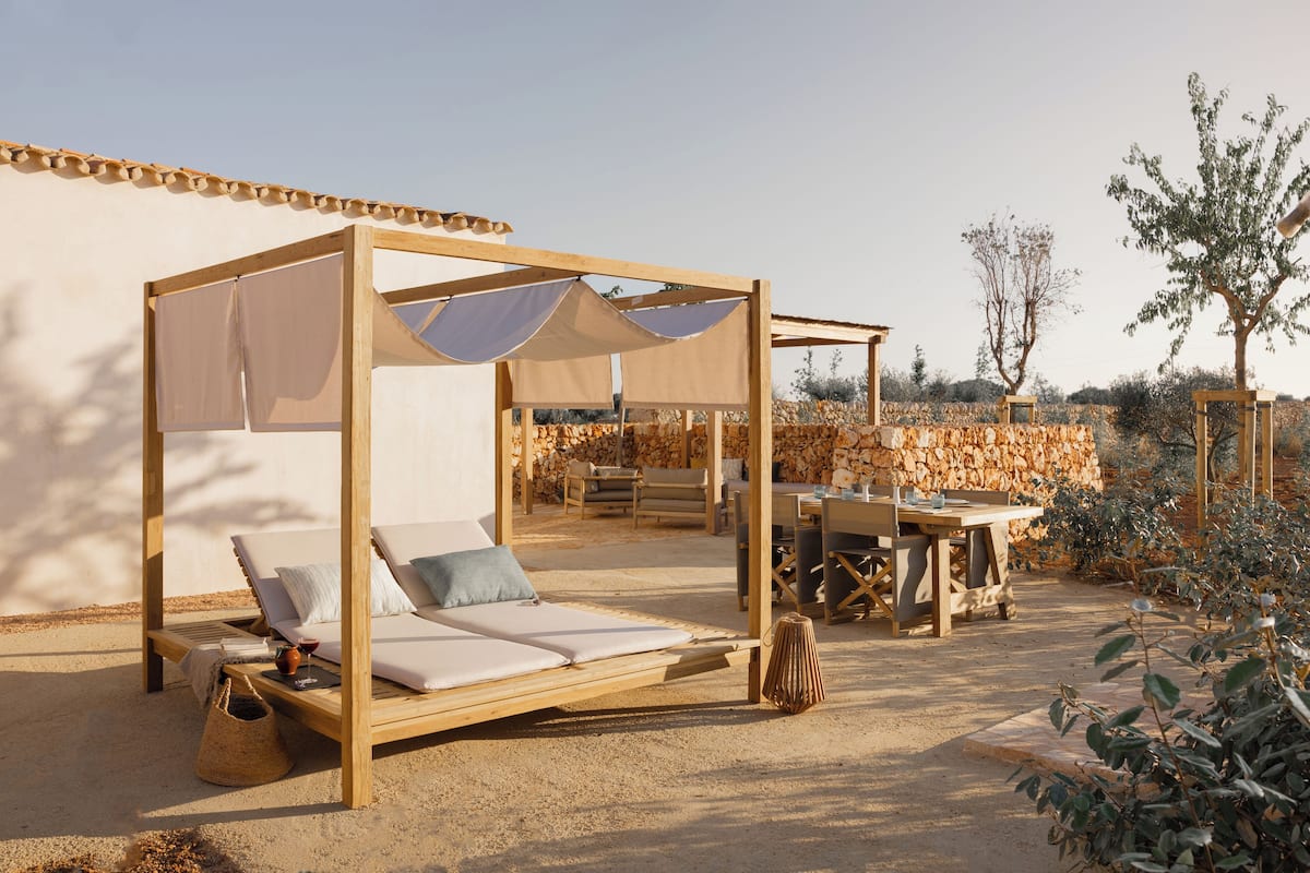 Una cama de día de madera con dosel, una mesa y sillas en un entorno exterior iluminado por el sol.