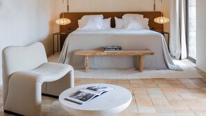 A bedroom with a wooden ceiling, a bed with white linens, a bedside table with a lamp, a chair, and a round table with a magazine.