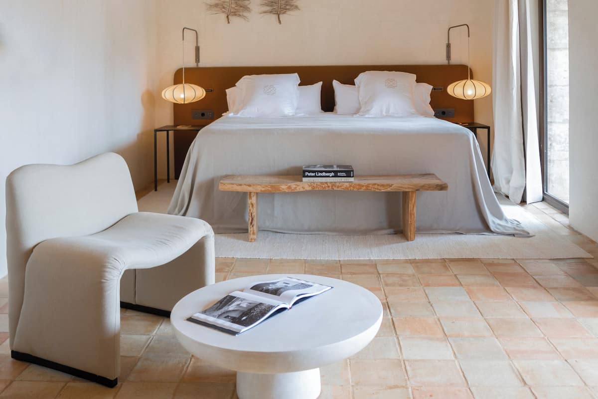 Un dormitorio con techo de madera, una cama con sábanas blancas, una mesita de noche con una lámpara, una silla y una mesa redonda con una revista.