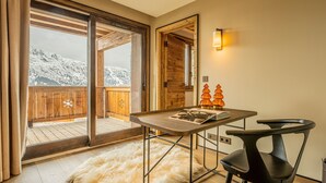 Une pièce avec une table en bois, une chaise noire, un tapis moelleux et une vue sur des montagnes enneigées, visible à travers une porte vitrée.