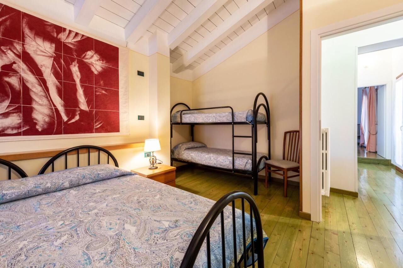 A room with a bunk bed, a single bed, a chair, a lamp, and a large abstract painting on the wall.