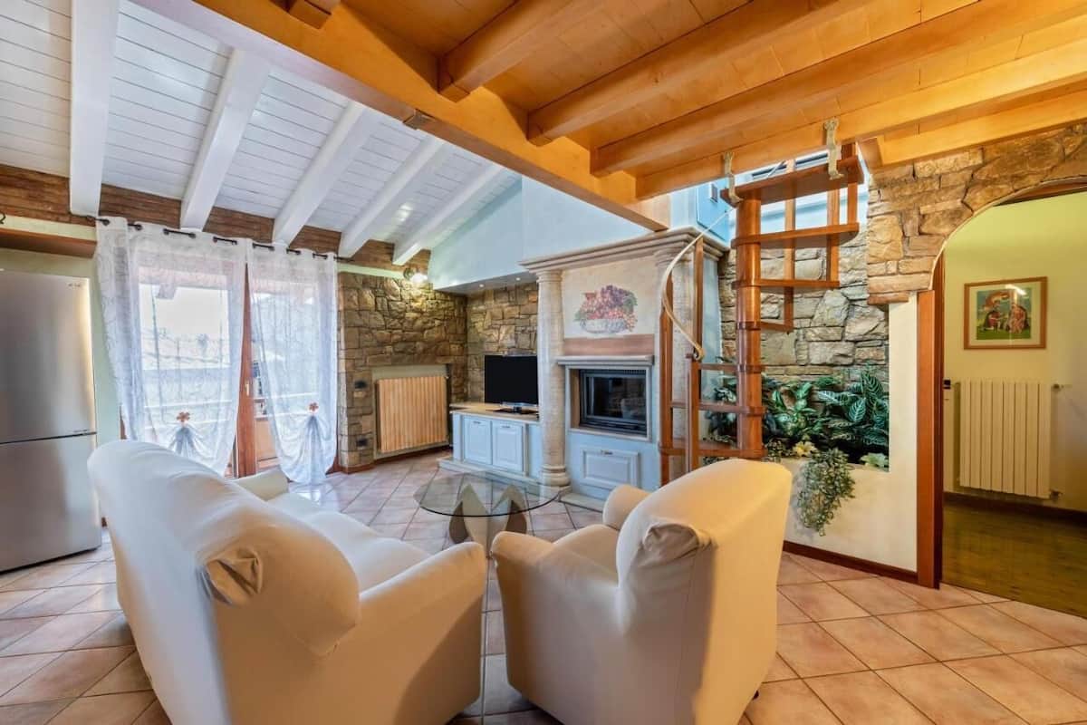 A cozy living room with a fireplace, stone walls, and a wooden ceiling. There are two white armchairs and a glass coffee table.