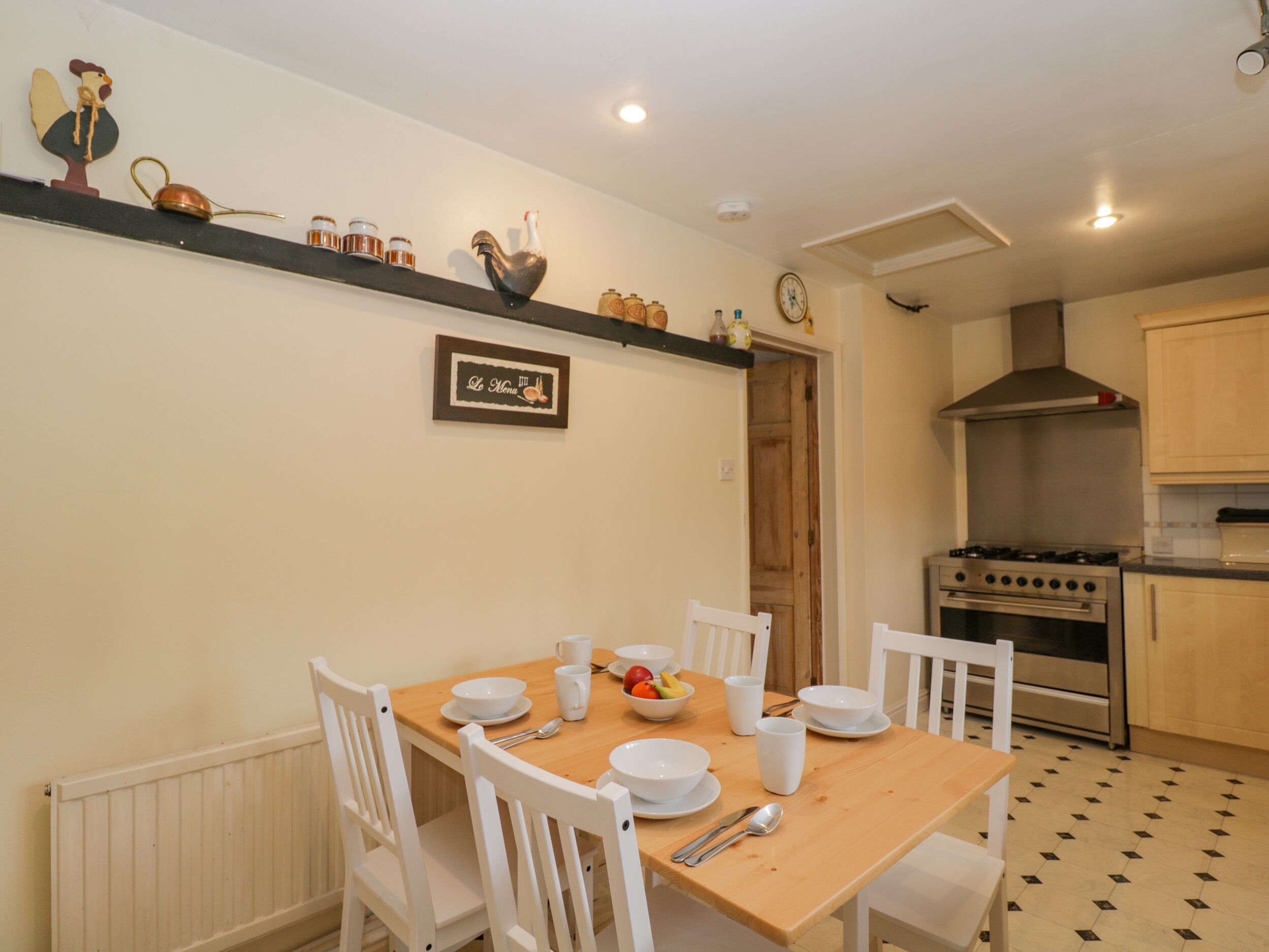 Uma sala de jantar com mesa de madeira e cadeiras brancas, uma cozinha com fogão e armários, e uma prateleira fixa com utensílios de cozinha.