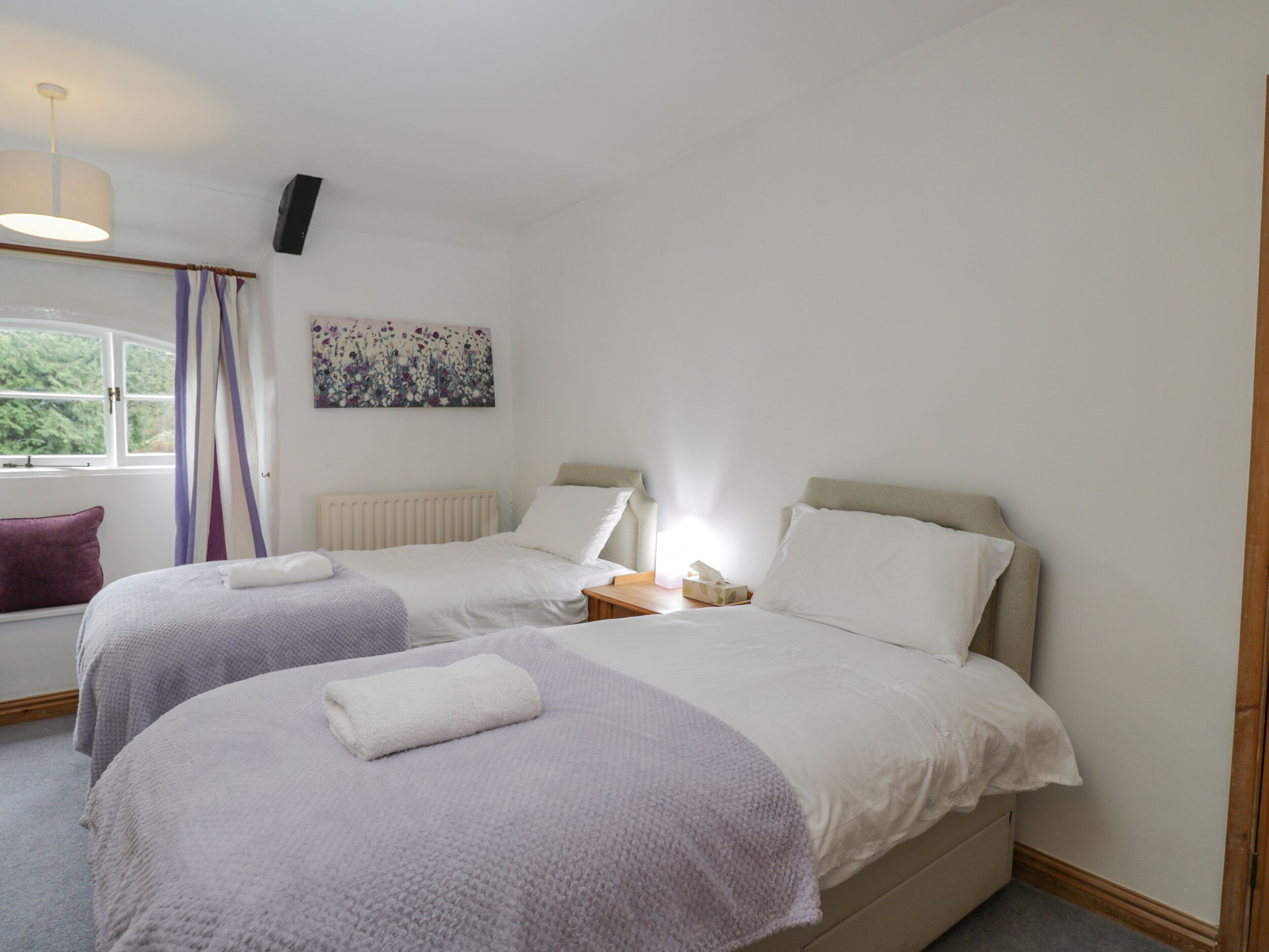 Une chambre d’hôtel avec deux lits, une fenêtre avec des rideaux, un radiateur, une table de chevet avec une lampe et un téléviseur fixé au mur.