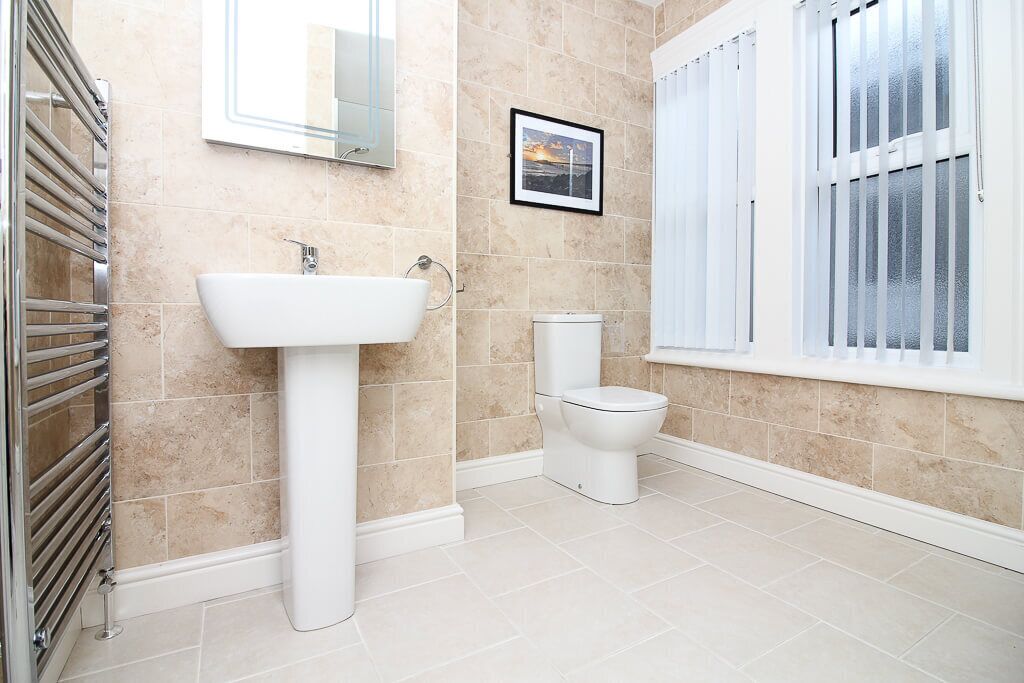 A bathroom with beige tiles, a white sink, a toilet, and a window with blinds.