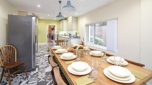 A dining area with a long wooden table set for four, a refrigerator, and a kitchen in the background. - The Beach House Luxury Rooms (Weston-super-Mare)