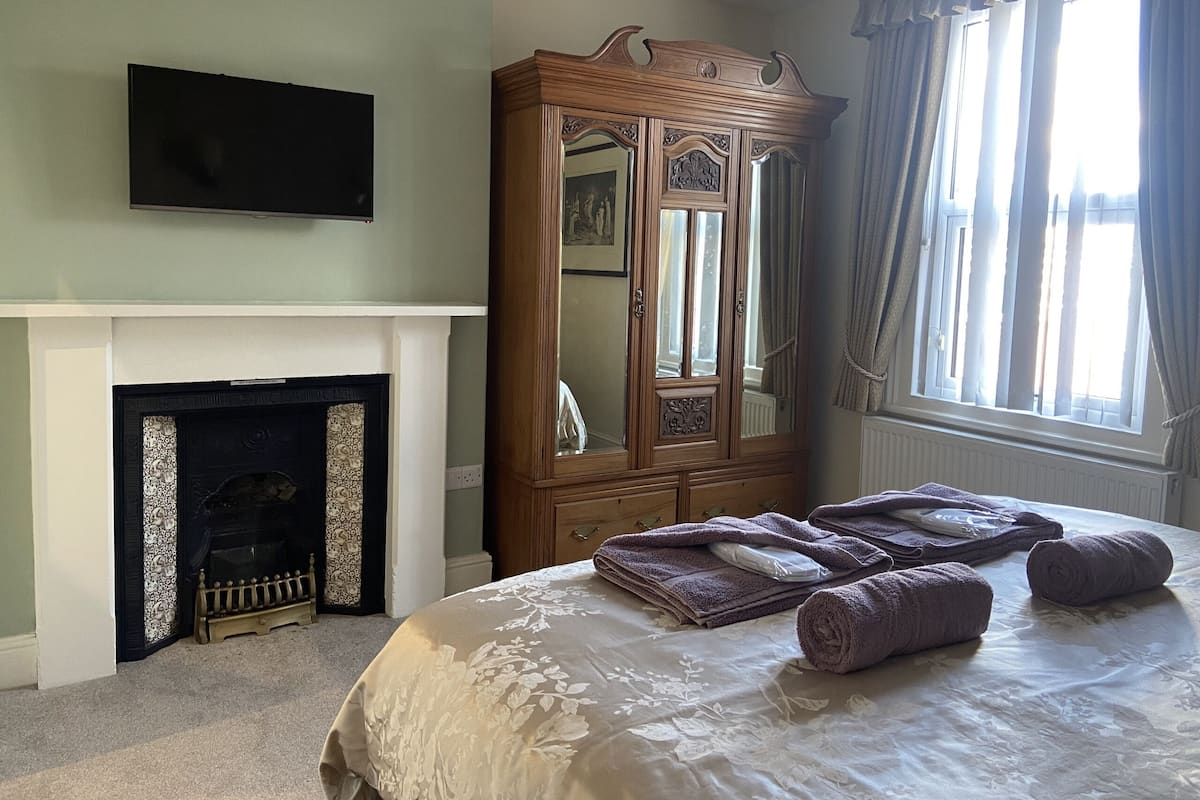A bedroom with a fireplace, a large wooden armoire, a bed with rolled towels, and a television mounted above the fireplace.