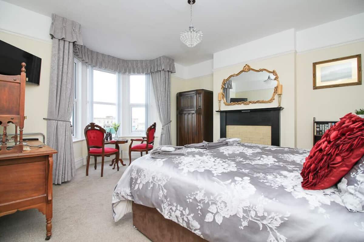 A bedroom with a large bed, a wooden dresser, a desk, two chairs, a fireplace, and a large mirror.