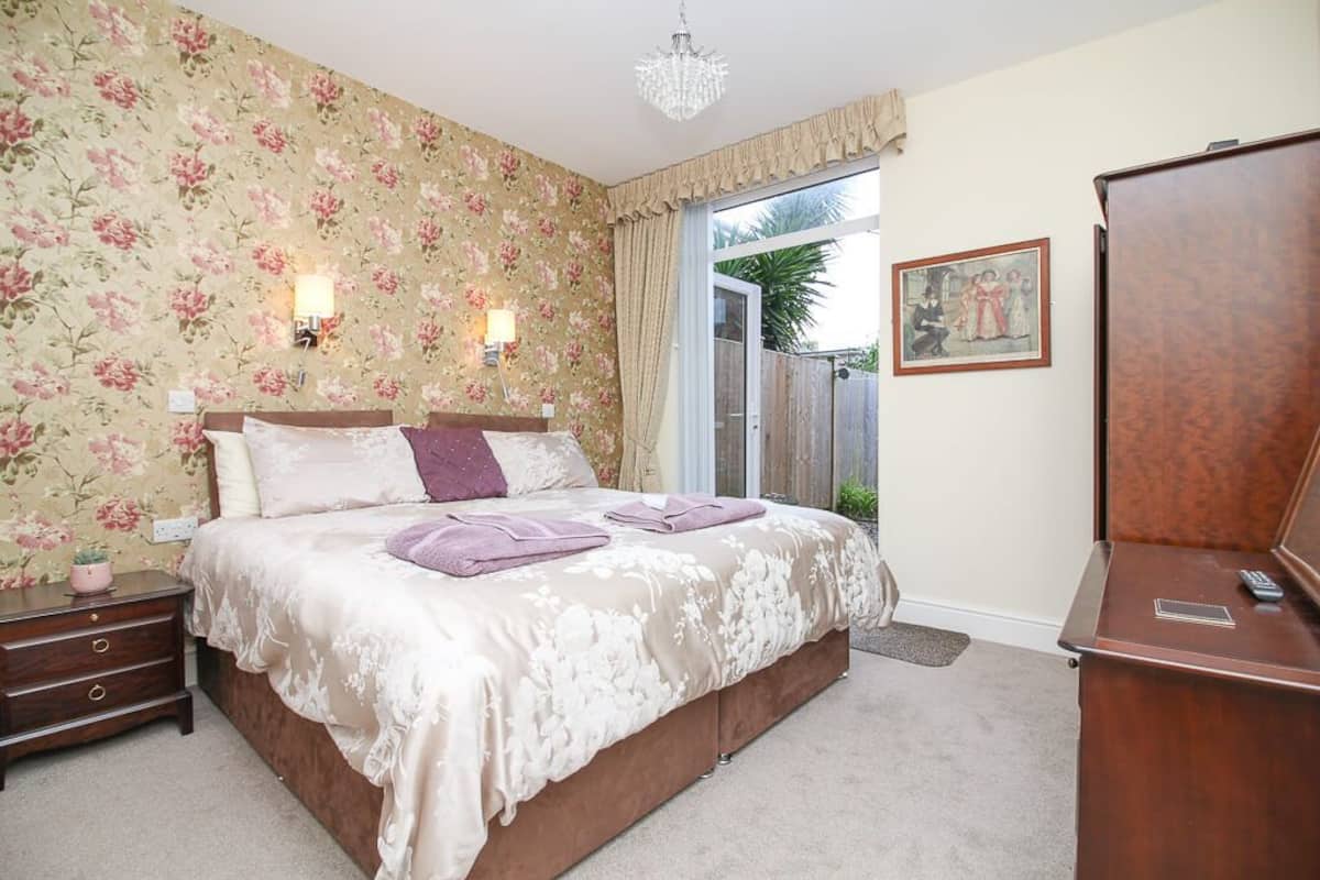 A bedroom with a floral wallpaper, a large bed, two bedside lamps, a wooden nightstand, a dresser, and a door leading outside.