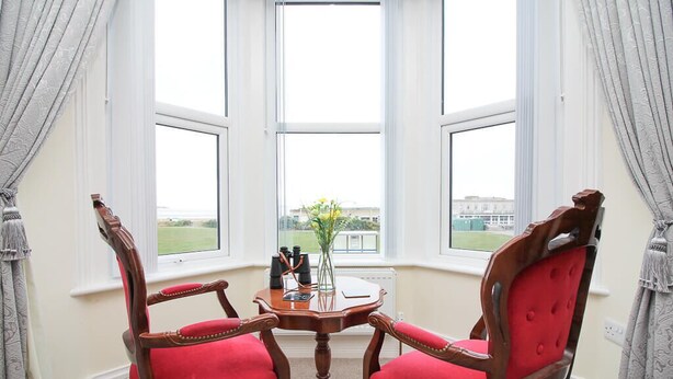 A room with a bay window, two red upholstered chairs, a small round table with a vase of flowers, and a view of a building across the street. - The Beach House Luxury Rooms (Weston-super-Mare)