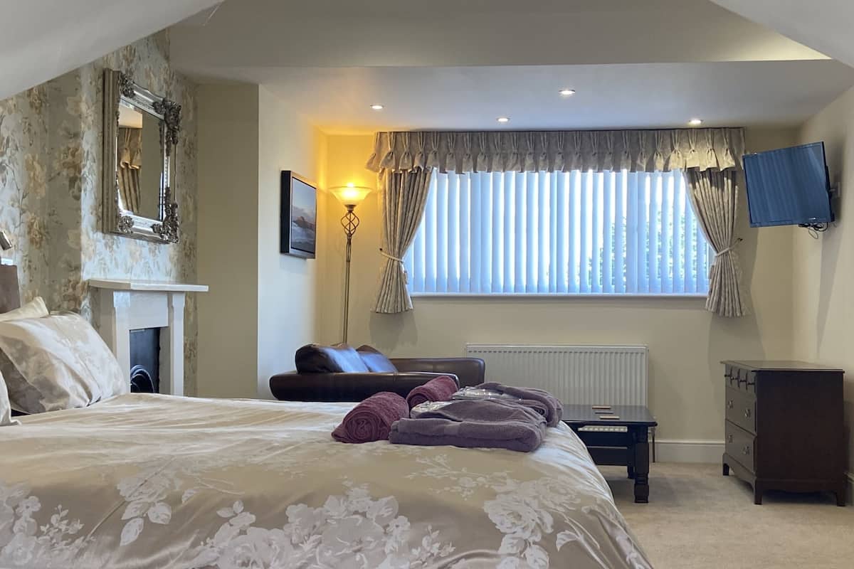 A bedroom with a large bed, a fireplace, a window with curtains, a desk, and a dresser.