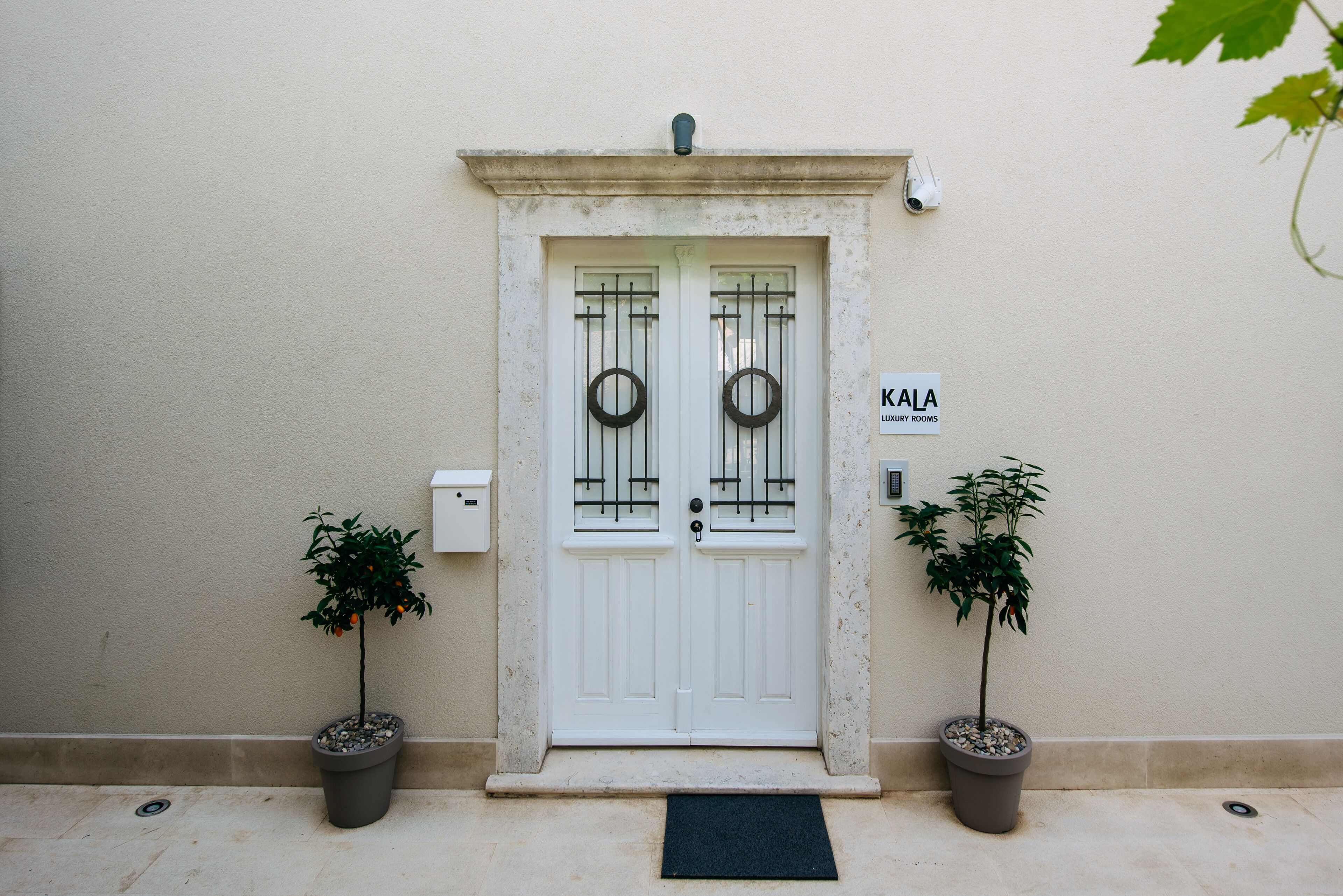 Eine weiße Tür mit einem Schild, auf dem 'KALA LUXURY ROOMS' steht, und je eine Topfpflanze auf beiden Seiten.