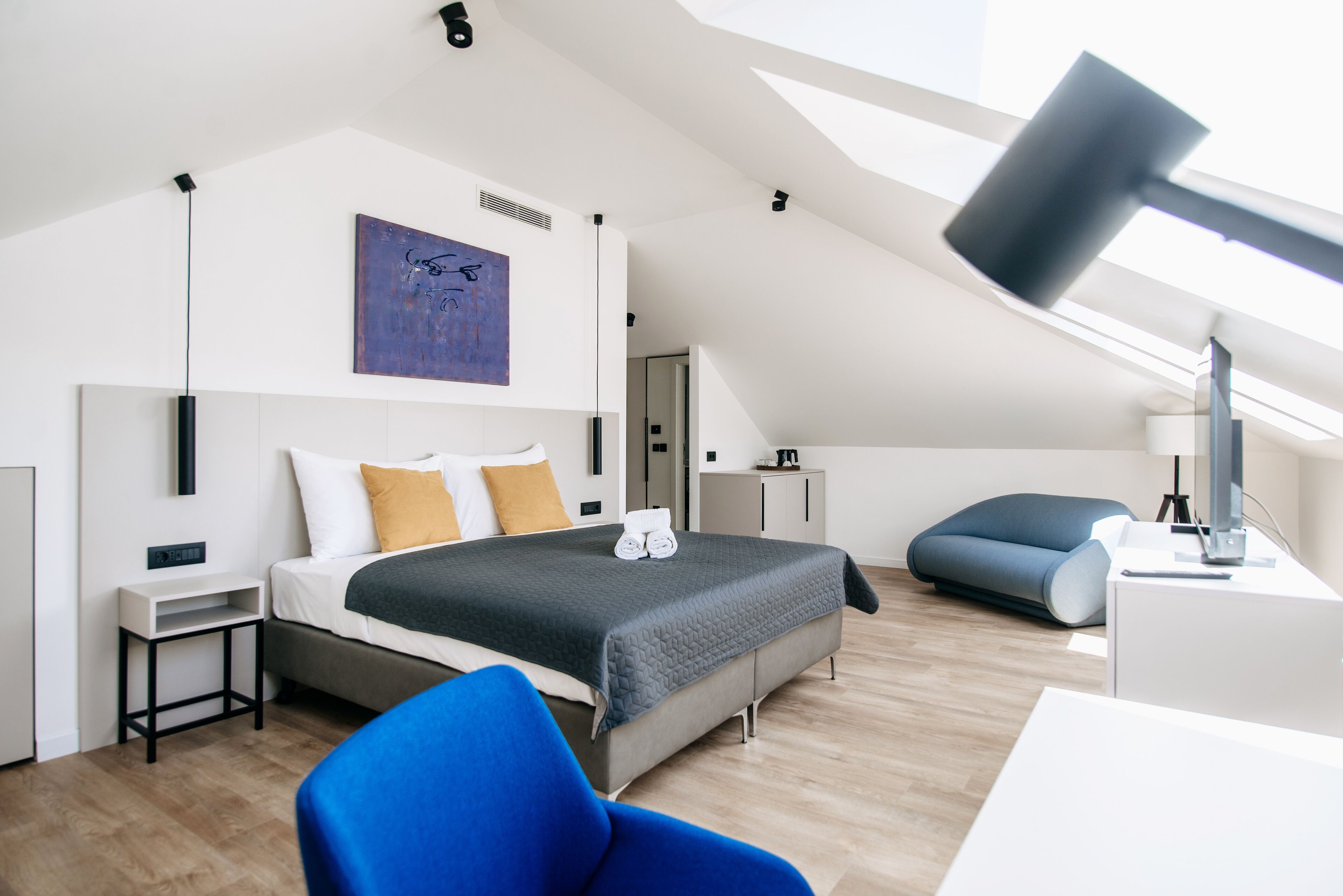 A modern bedroom with a sloped ceiling, a large bed with a grey and white color scheme, a blue chair, and a desk with a lamp.