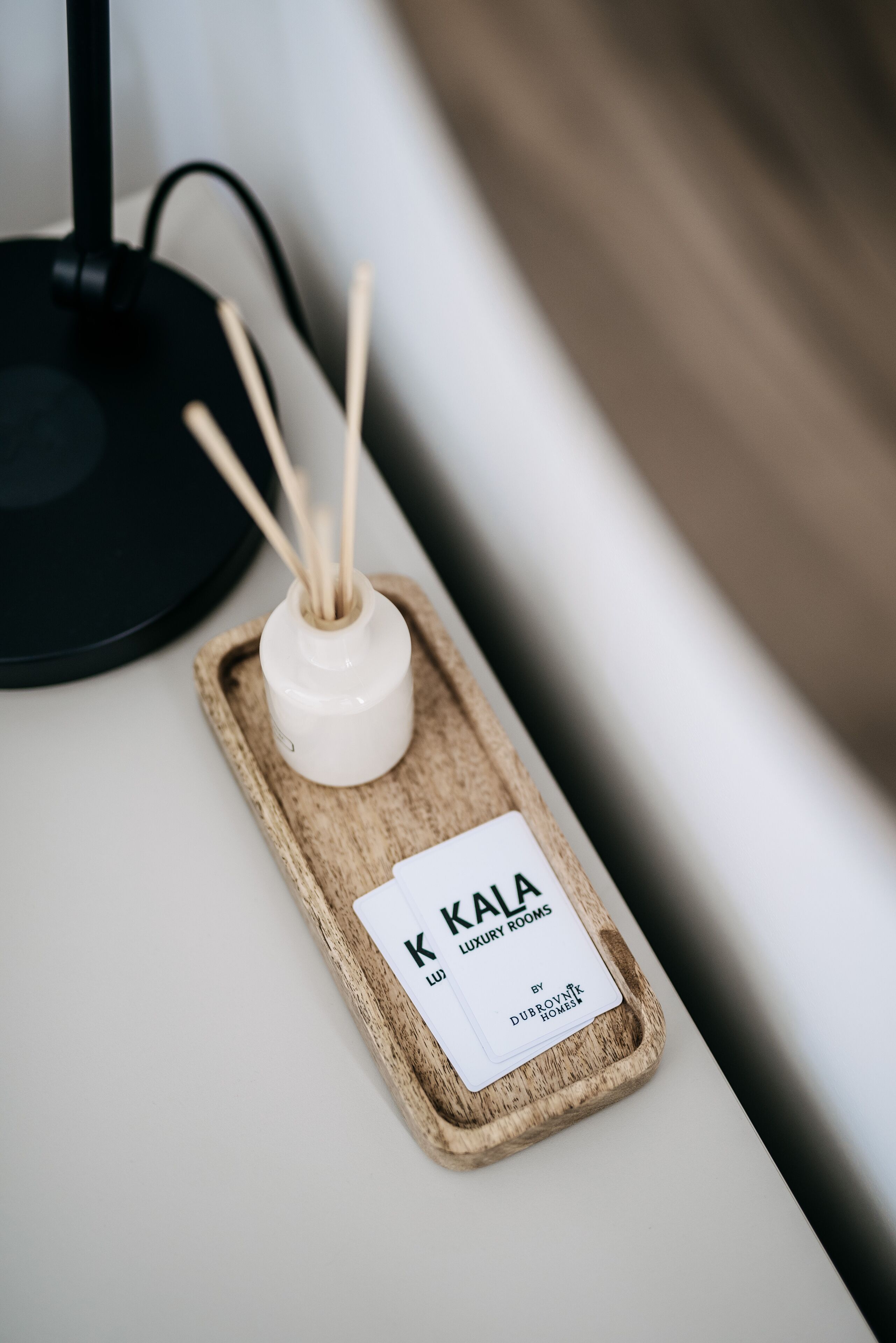 A white bottle with reed sticks on a wooden tray with a label reading "KALA LUXURY ROOMS by Dubrova".