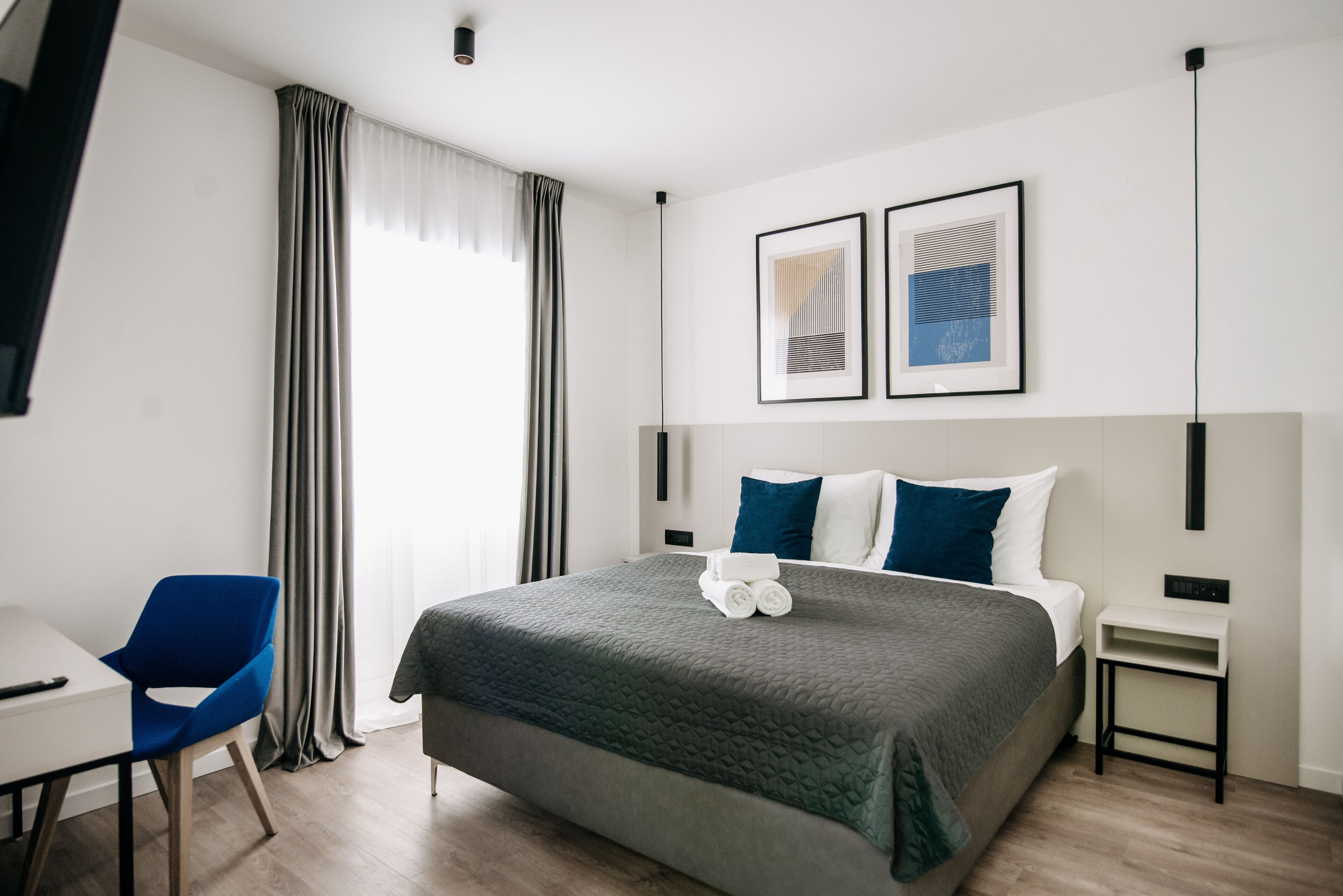 A modern hotel room with a large bed, a desk, and a chair. There are two framed artworks on the wall and a bedside table with a lamp.
