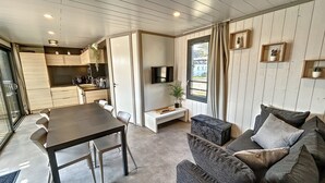 A modern living room with a dark sofa, a dining table, and a kitchen in the background. - Cozy Chalet for 6 with Heated Pool, Terrace & Parking, Near Beach (Jullouville)