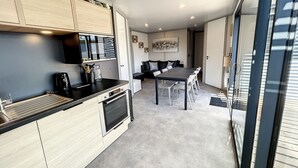 A modern kitchen with a dining area, featuring a large table and chairs, a coffee machine, and a built-in oven. - Cozy Chalet for 6 with Heated Pool, Terrace & Parking, Near Beach (Jullouville)