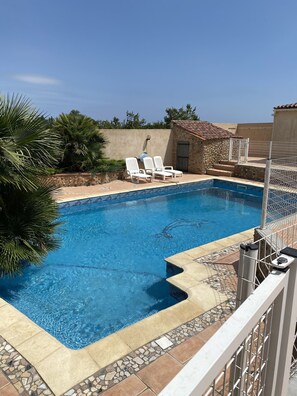 Une piscine à l’eau d’un bleu cristallin, bordée de pierres et de carreaux. On y trouve des chaises longues blanches et, au fond, un petit bâtiment en pierre.