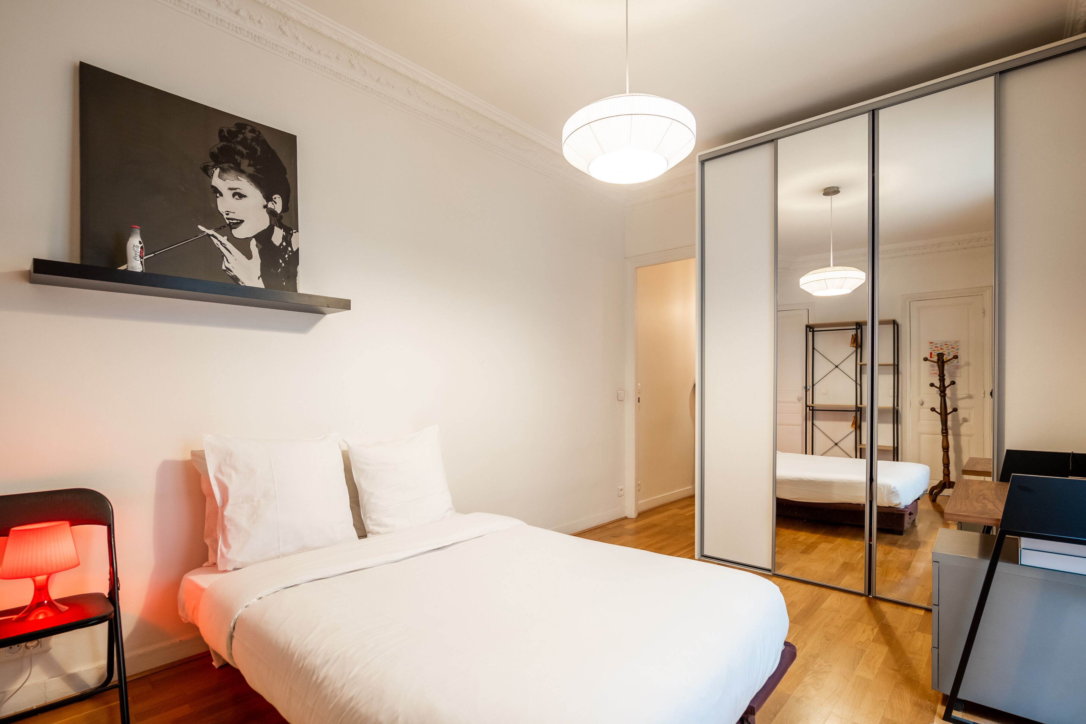 A modern bedroom with a bed, bedside table, and a red lamp. There is a painting on the wall and a sliding door leading to another room.