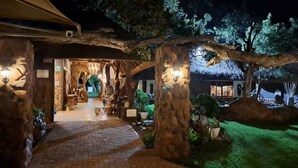A well-lit pathway leading to a building with a rustic design, surrounded by trees and potted plants. - Kilima Private Game Reserve and Spa (Kareeberg)
