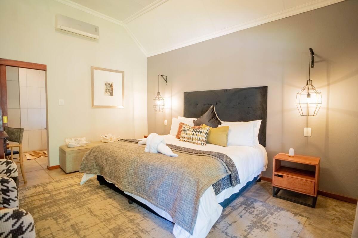 A hotel room with a large bed, a bedside table, a nightstand, a chair, and a framed picture on the wall.