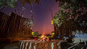 A stone fire pit with logs, surrounded by a wooden fence and trees, under a twilight sky. - Kilima Private Game Reserve and Spa (Kareeberg)