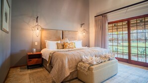 A bedroom with a large bed, a nightstand, a bench, and a view of the outdoors through a large window. - Kilima Private Game Reserve and Spa (Kareeberg)