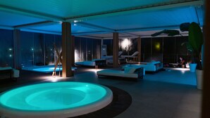 A modern indoor pool area with a circular hot tub, lounge chairs, and a view of the city at night. - Laghi Nabi (Castel Volturno)