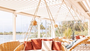 A spacious, sunlit room with large windows overlooking a body of water, featuring wicker furniture and a hanging chair. - Laghi Nabi (Castel Volturno)