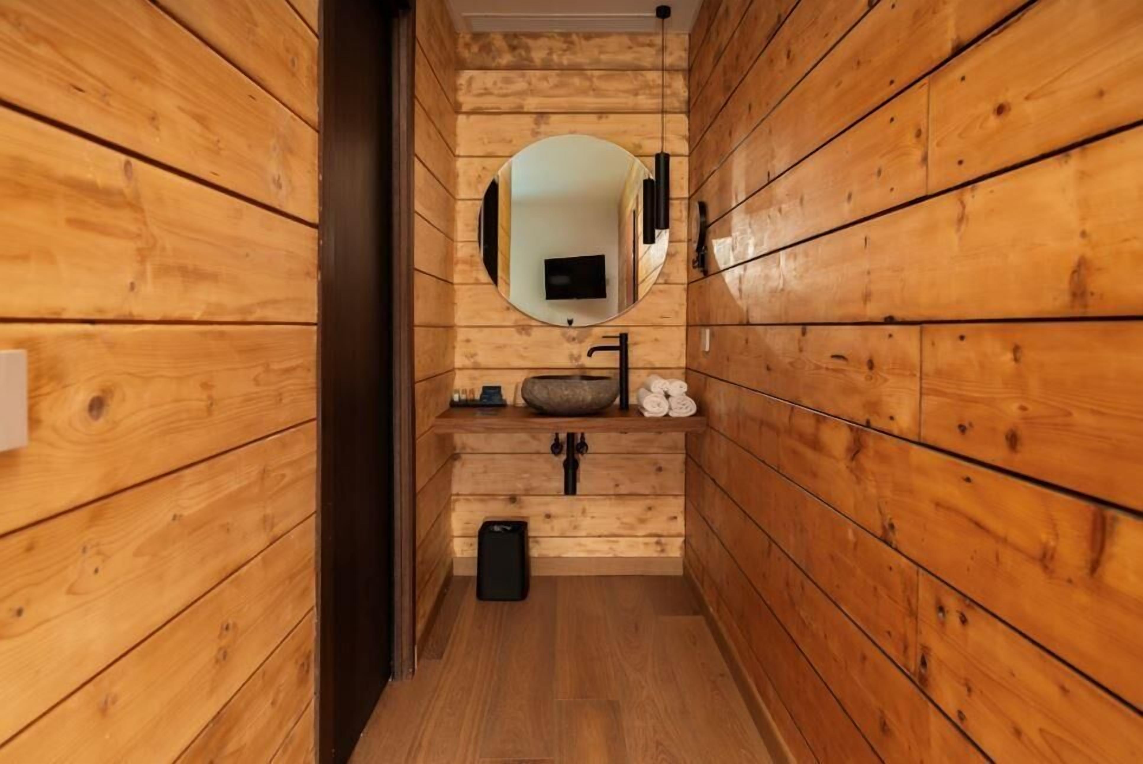 Interno di una baita di legno con parete in legno, uno specchio rotondo, un lavandino e un cestino nero.