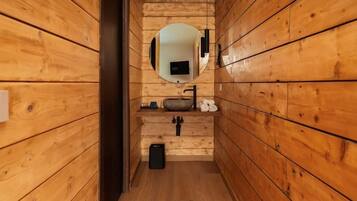 Interno di una baita di legno con parete in legno, uno specchio rotondo, un lavandino e un cestino nero.