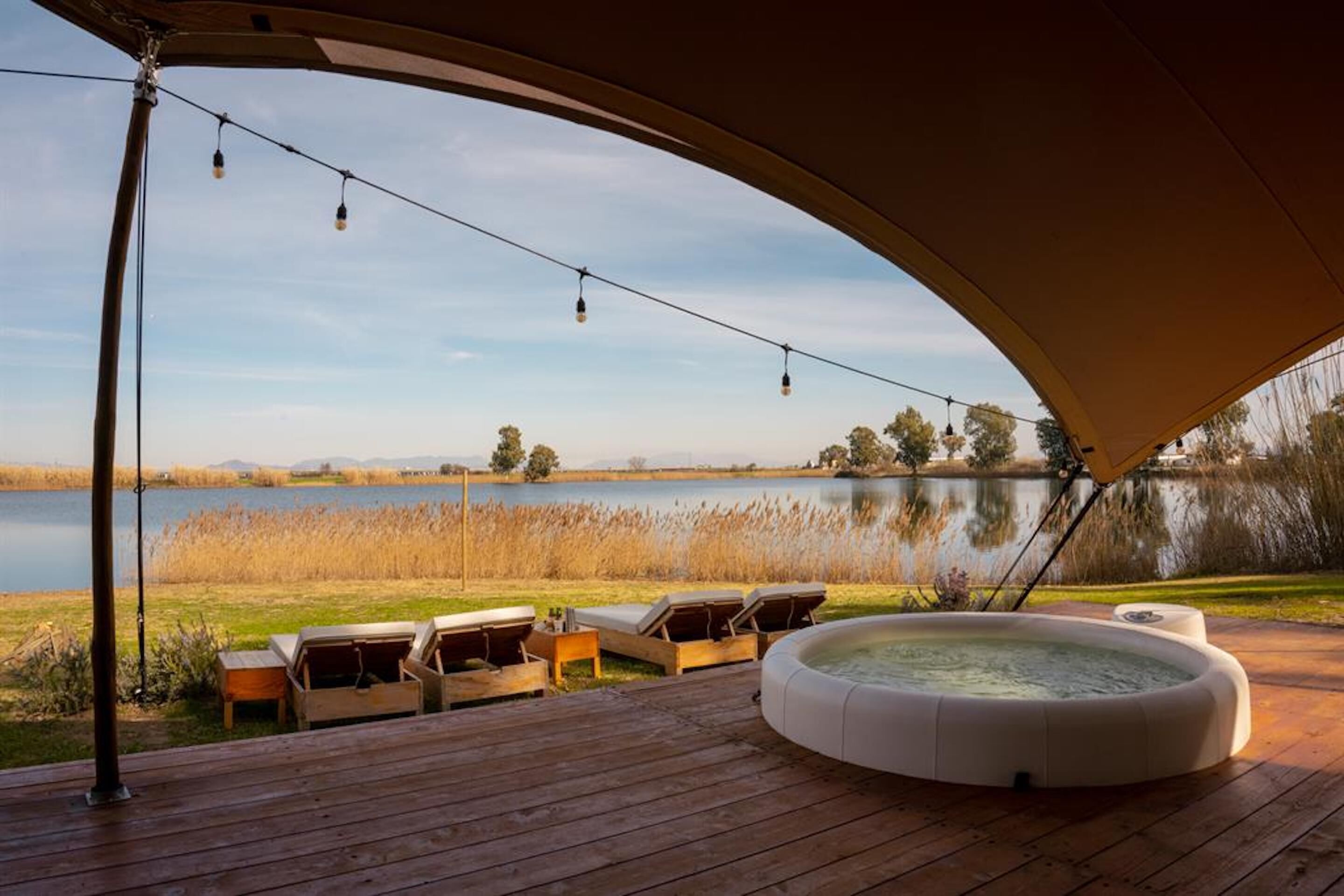 Terraza exterior con sillones de descanso, jacuzzi y luces colgantes sobre un lago.