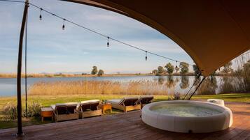 Terraza exterior con sillones de descanso, jacuzzi y luces colgantes sobre un lago.