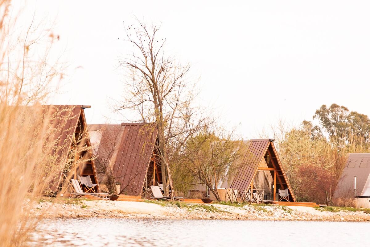 Une rangée de cabanes en bois rustiques, situées au bord d’un plan d’eau, chacune dotée d’une petite terrasse et d’un espace salon.