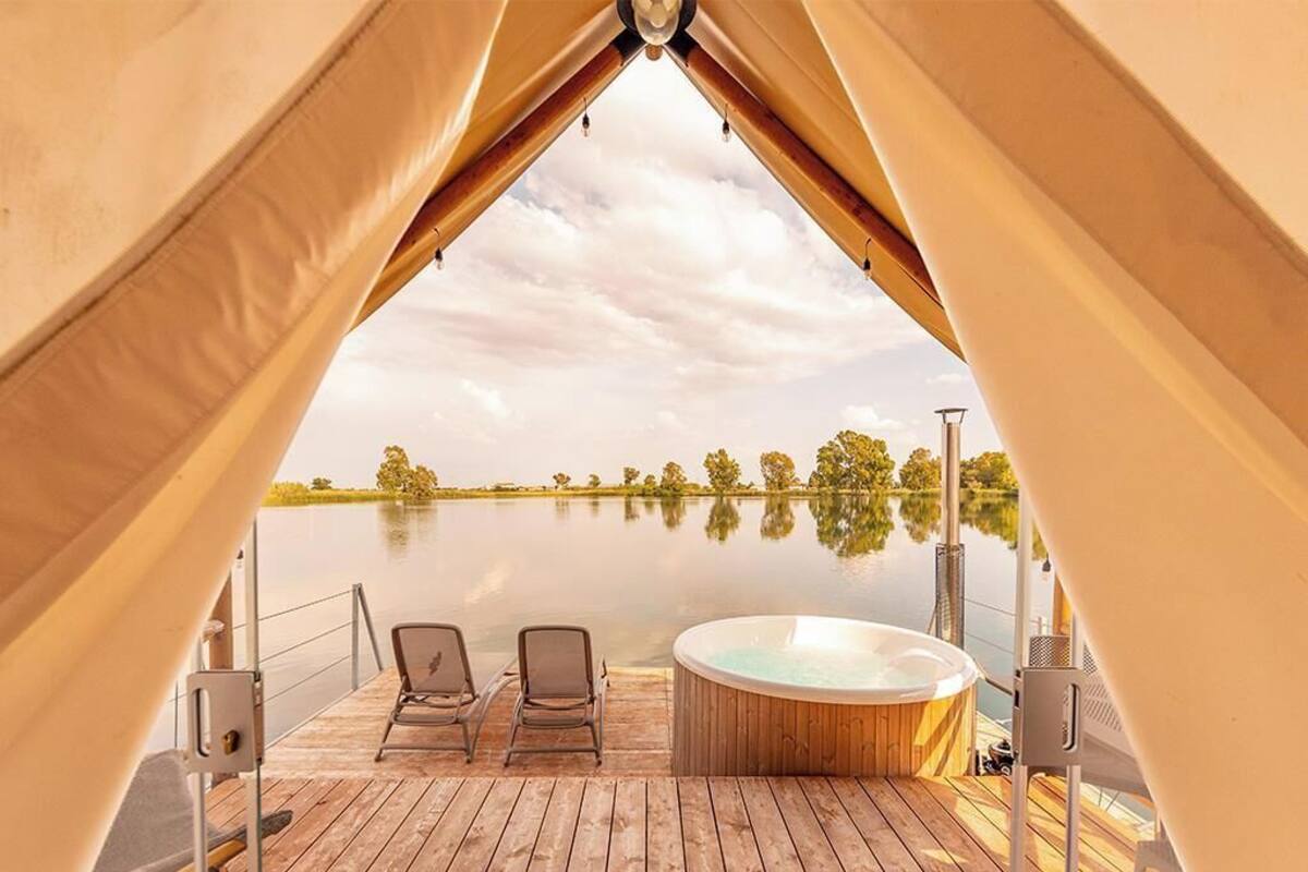 Un espace aménagé sous une tente, avec un jacuzzi et deux fauteuils donnant sur un lac paisible.