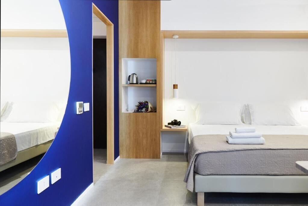 Una habitación de hotel moderna con una pared azul curva, un mueble de madera, una cama con manta gris y una mesita de noche con lámpara.