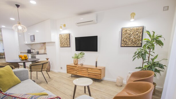 Una sala de estar moderna con un sofá gris, una mesita redonda pequeña, un mueble de madera y dos sillas marrones. Un televisor de pantalla plana está montado en la pared sobre el mueble.
