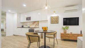 Una cocina moderna con armarios blancos, encimera de mármol, una mesa redonda con dos sillas, un televisor montado en la pared y un mueble de madera con plantas decorativas.