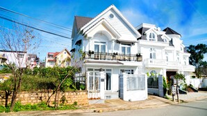 A large, white two-story house with a balcony, a gated entrance, and a stone wall in the foreground. - Mon Bac Home (Lam Vien - Da Lat)