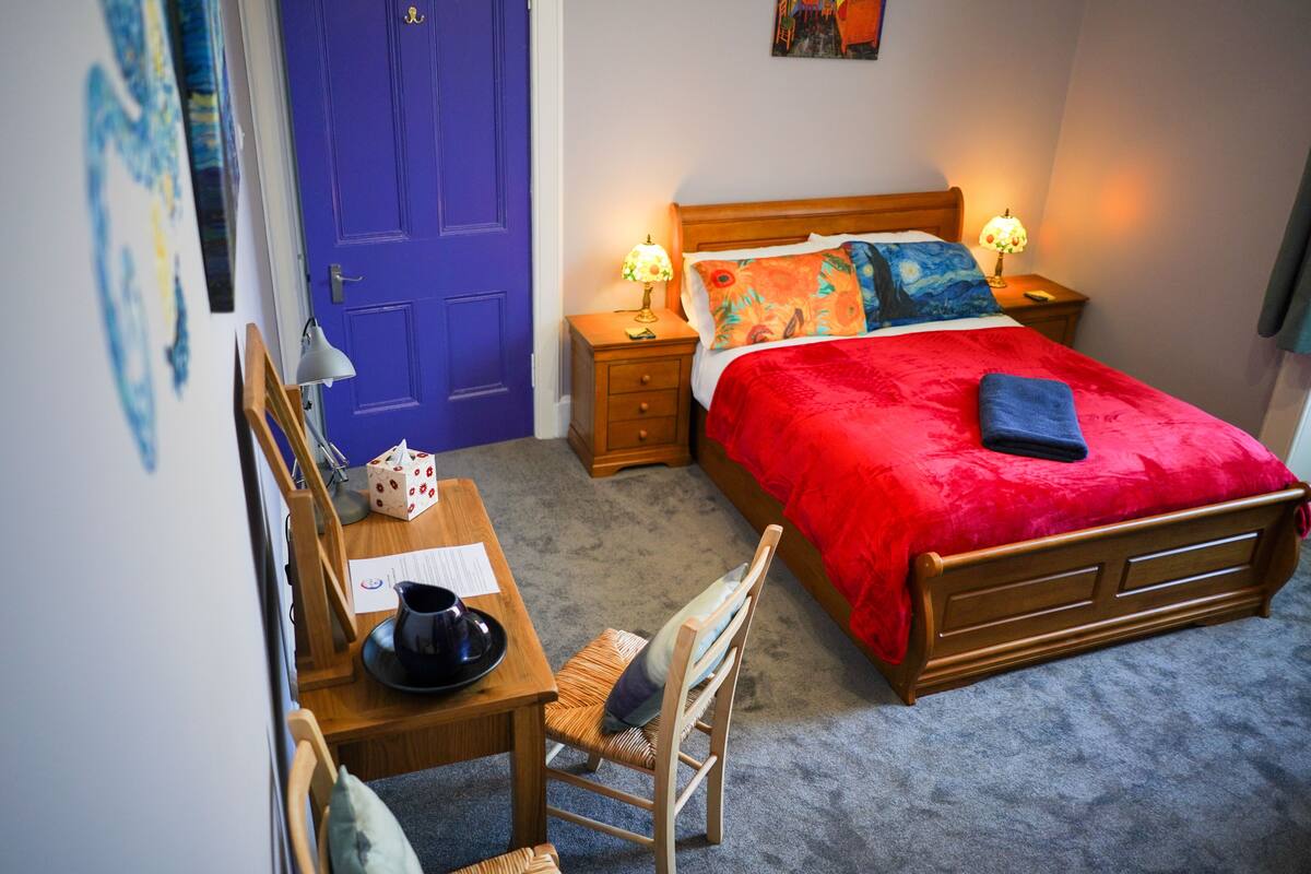 A bedroom with a wooden bed, red and blue bedding, two bedside lamps, a wooden nightstand, a desk with a chair, and a blue door.