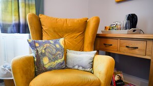 A yellow armchair with a colorful pillow, a wooden desk with a coffee maker, and a shelf with books. - The Art Bank (Dalbeattie)